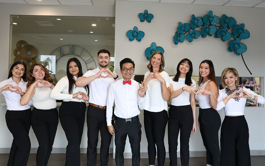 A group of eight people, dressed in business casual attire, stand in a row indoors, smiling and making heart shapes with their hands. Blue butterfly wall art is visible behind them.